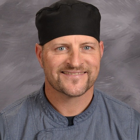 A man wearing a chef's hat and a black shirt, standing confidently in a kitchen setting.