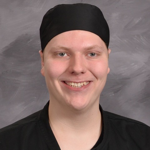 A young man wearing a grey shirt and apron, standing confidently with a friendly expression.