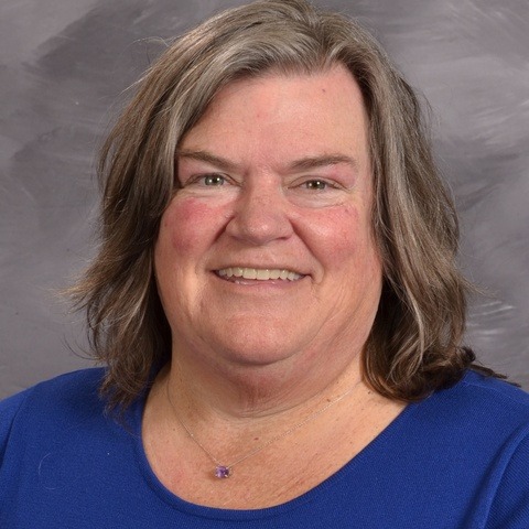 A woman with long hair is wearing a gray cardigan, standing in a relaxed pose against a neutral background.