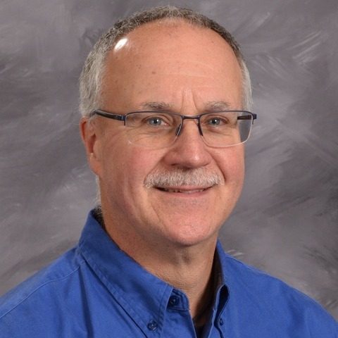A man in a blue shirt and tie stands confidently in front of a gray wall, exuding professionalism and poise.