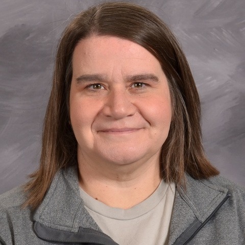 A woman with long hair wearing a blue lanyard, standing confidently with a neutral background.