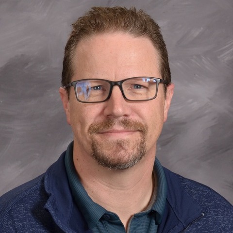 A man wearing a blue sweatshirt smiles warmly at the camera, exuding a friendly and approachable demeanor.