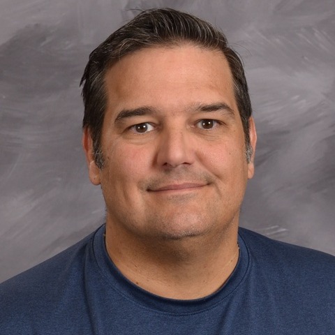 A man wearing a black vest over a green shirt, standing confidently with a neutral expression.