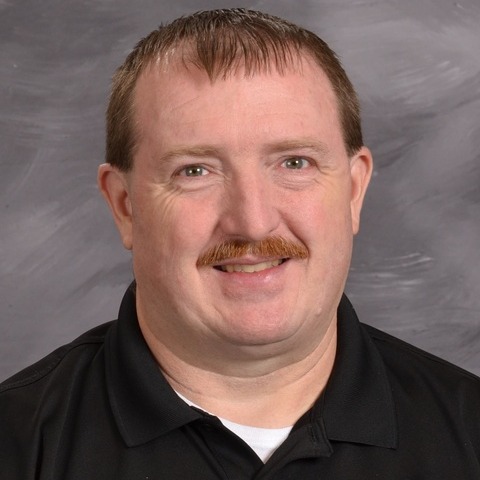 A bearded man wearing a blue shirt, standing confidently with a neutral expression.