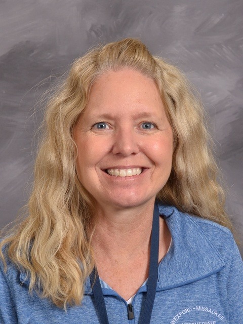 A woman with long blonde hair is wearing a blue shirt, standing confidently with a pleasant expression.