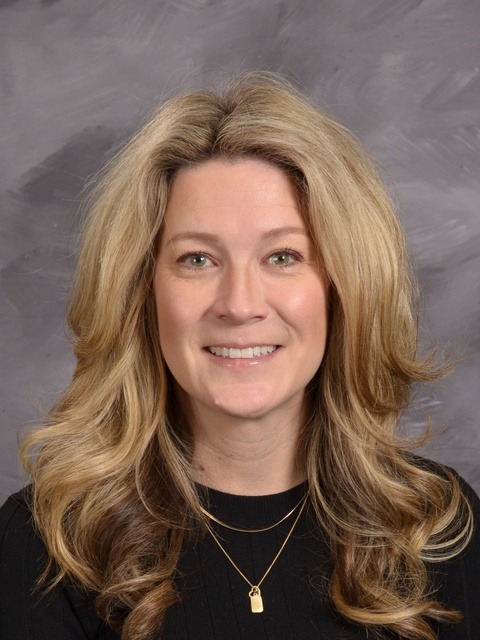  A woman with long hair wearing a tan cardigan, standing confidently with a warm smile.