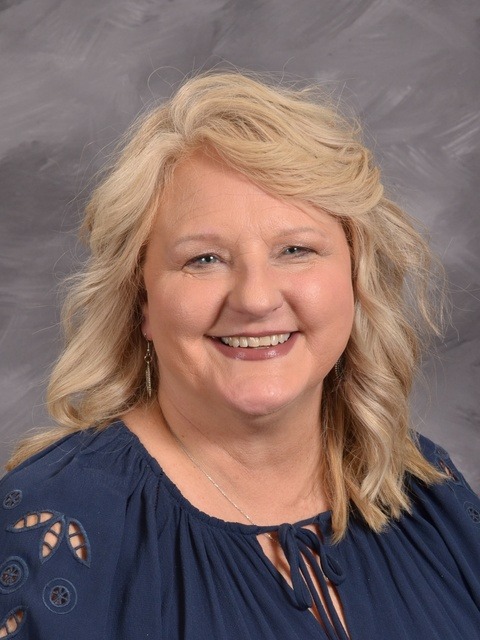 A woman with blonde hair wearing a blue shirt, smiling and standing against a neutral background.