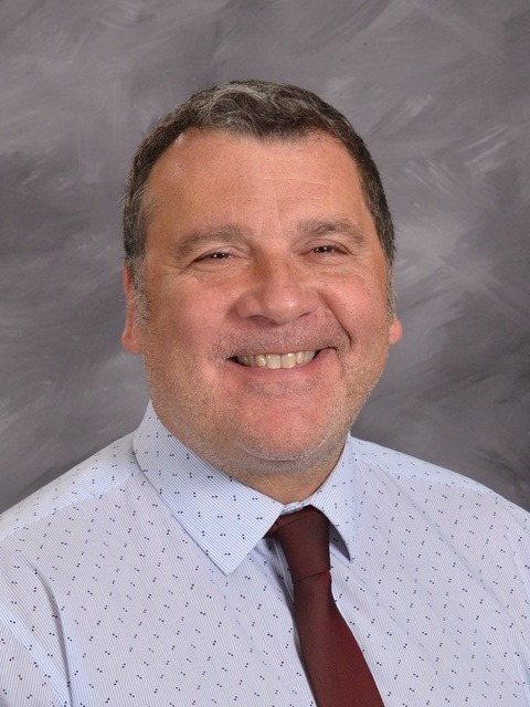 A man wearing a blue shirt and a green tie smiles warmly at the camera, exuding a friendly demeanor.