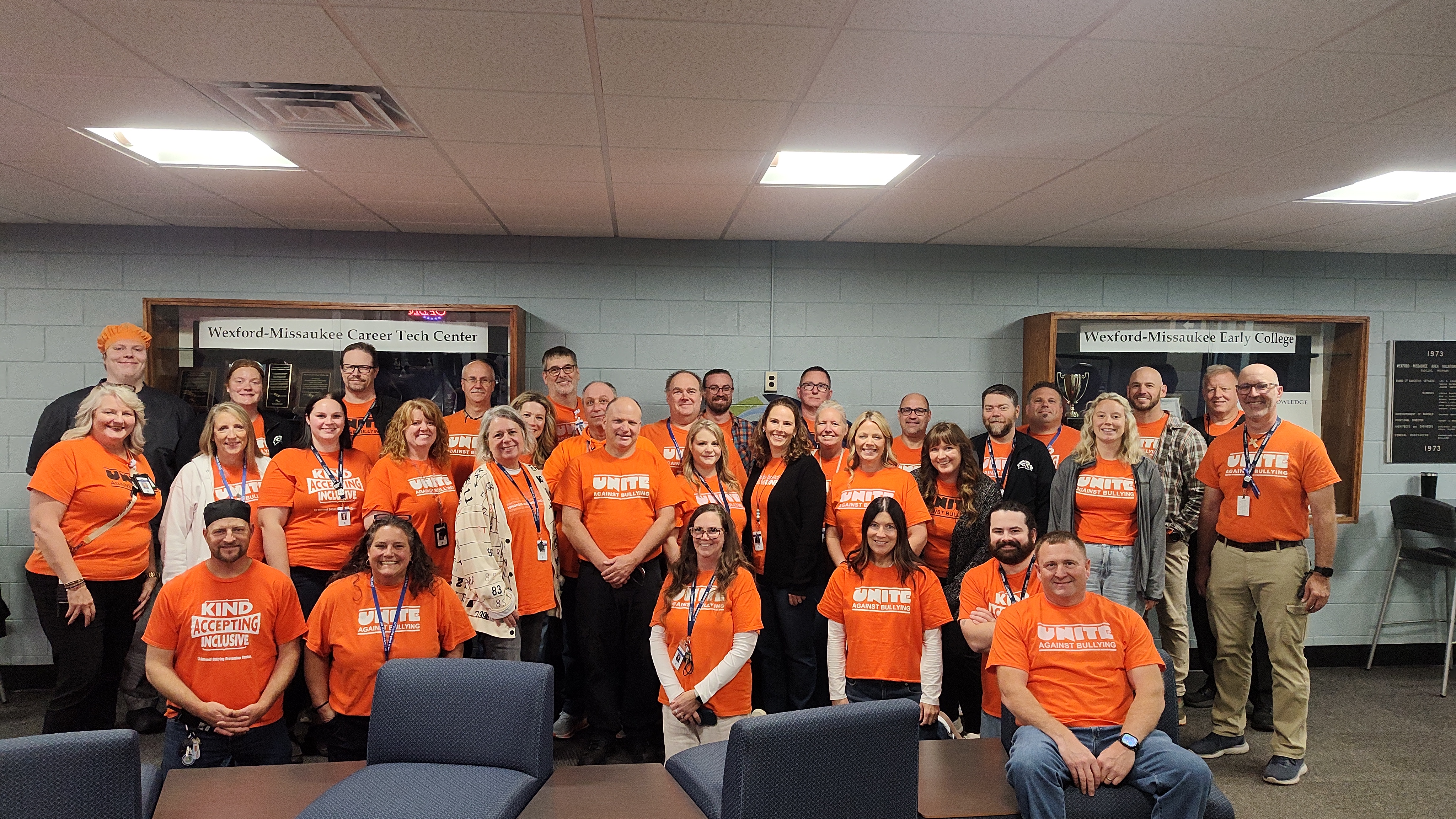 This image shows a large group of people posing together indoors, likely for an anti-bullying campaign. Most of the individuals are wearing bright orange t-shirts with the words "UNITE Against Bullying" or "KIND ACCEPTING INCLUSIVE" printed on them. In the background, there are display cases with signs that read "Wexford-Missaukee Career Tech Center" and "Wexford-Missaukee Early College," suggesting the photo was taken at an educational facility.
