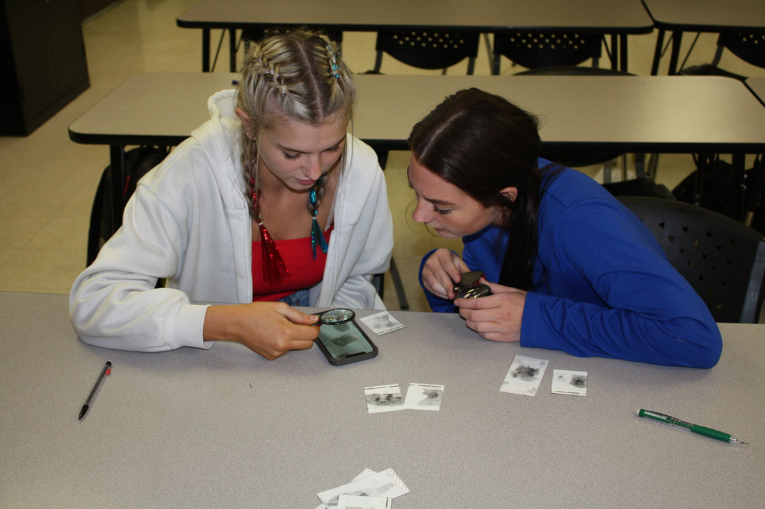 Two young women are closely examining photographs on a smartphone while sitting at a table, with additional photos laid out in front of them.