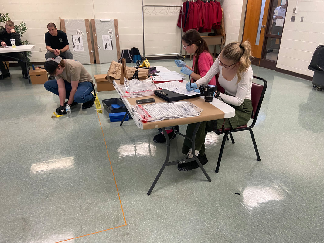 A group of individuals is engaged in an activity in a room, with two people seated at a table working with materials, while another person is on the floor measuring or examining something.