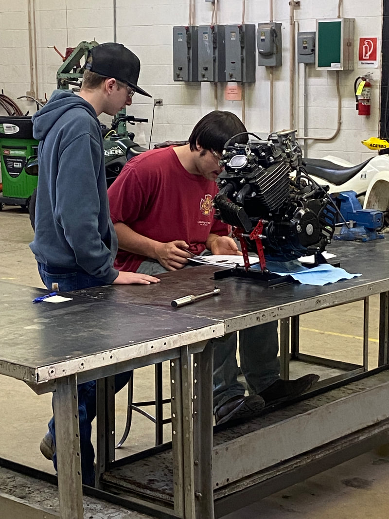 Two individuals are working together at a metal table in a workshop, focusing on an engine assembly while surrounded by various tools and equipment.