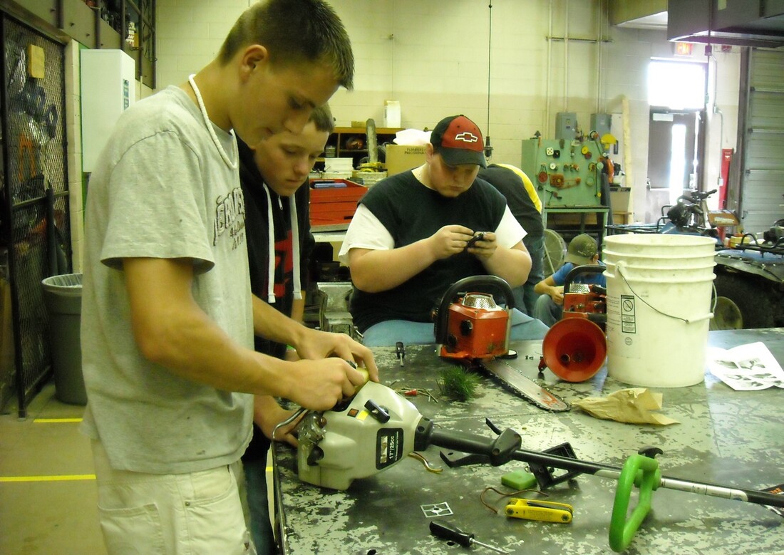 Three students are working on small engines at a workshop table, with tools and equipment scattered around them.