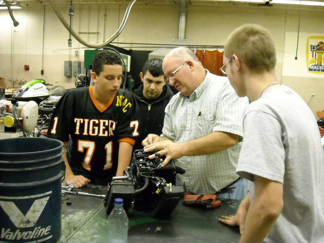 A group of four individuals, including a teacher and three students, are gathered around a workbench in a workshop, examining a mechanical component together.