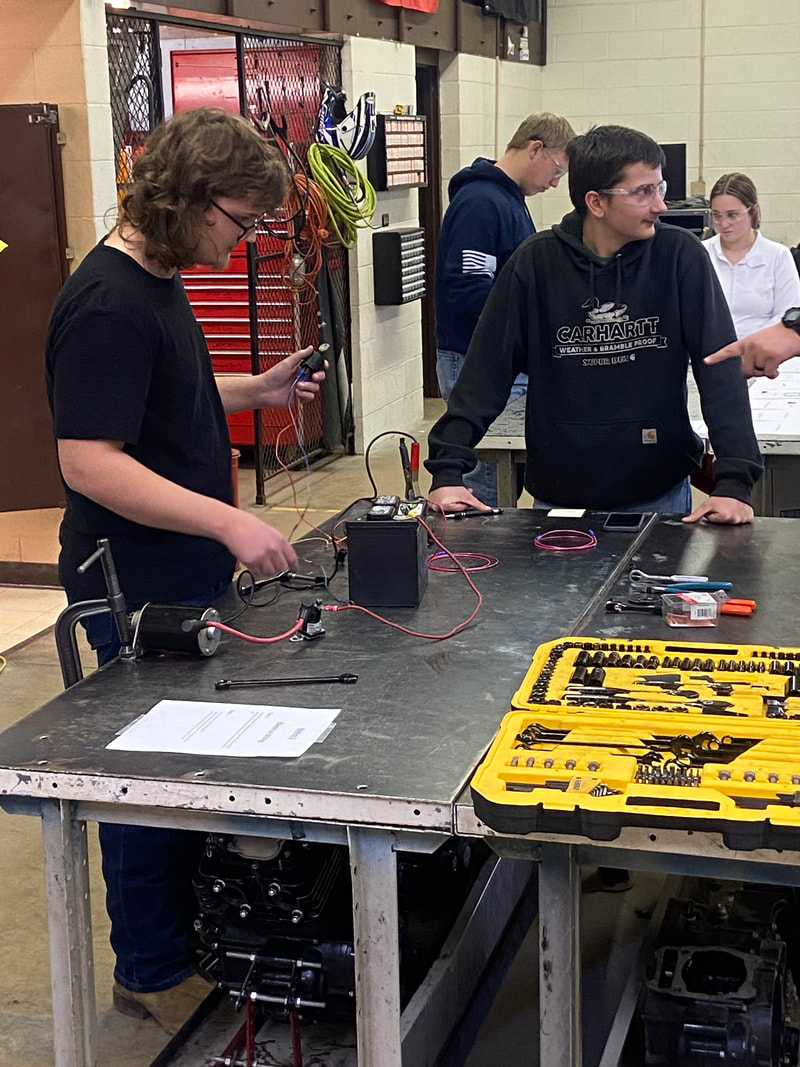Students are engaged in a hands-on electrical project at a workbench, with tools and equipment visible around them.