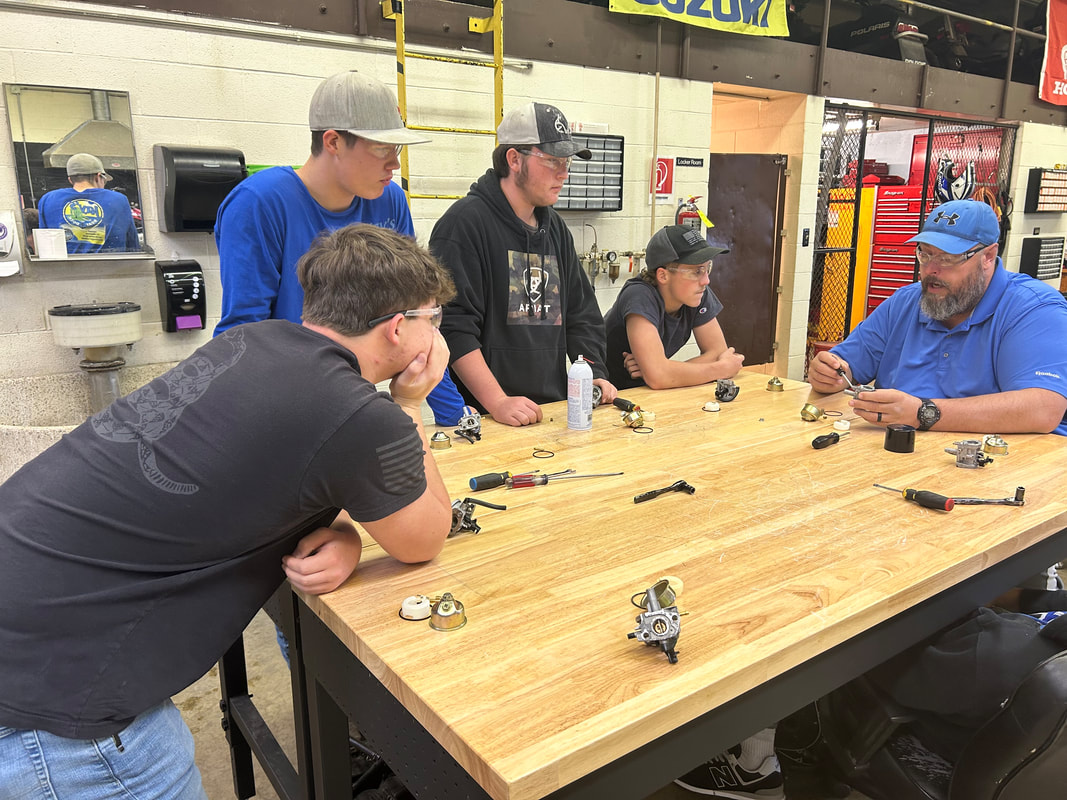 A group of five individuals, including a teacher and four students, are gathered around a wooden table in a workshop, engaged in a hands-on learning activity involving mechanical parts and tools.