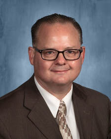 A man in a suit and tie stands confidently, smiling for the camera in a professional setting.
