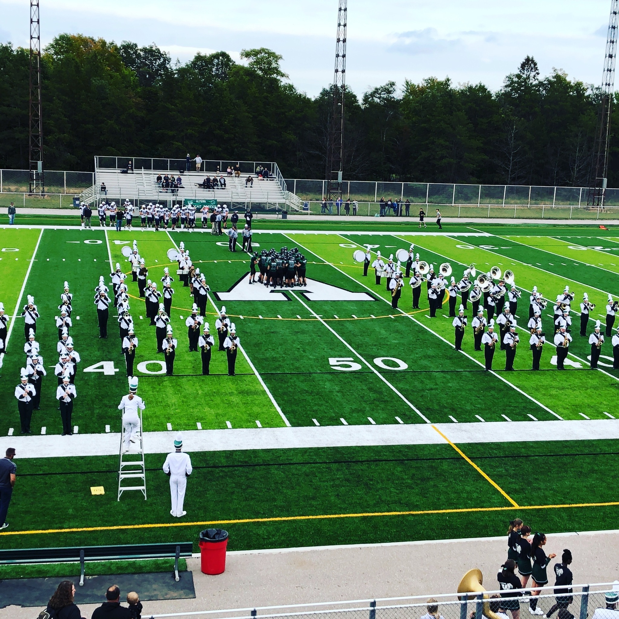 A marching band performs on a football field, with musicians arranged in formation and spectators in the background.