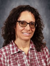 A woman with dark, curly hair wearing a red and white plaid shirt against a soft gray backdrop, exuding a friendly demeanor.