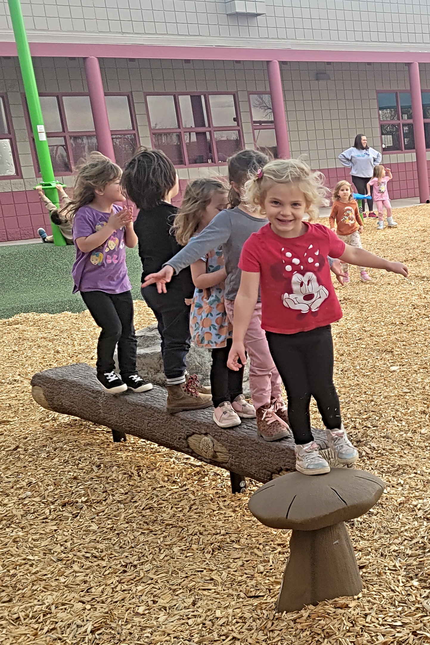 A group of children playfully balances on a log at an outdoor playground, surrounded by wood chips and colorful equipment.