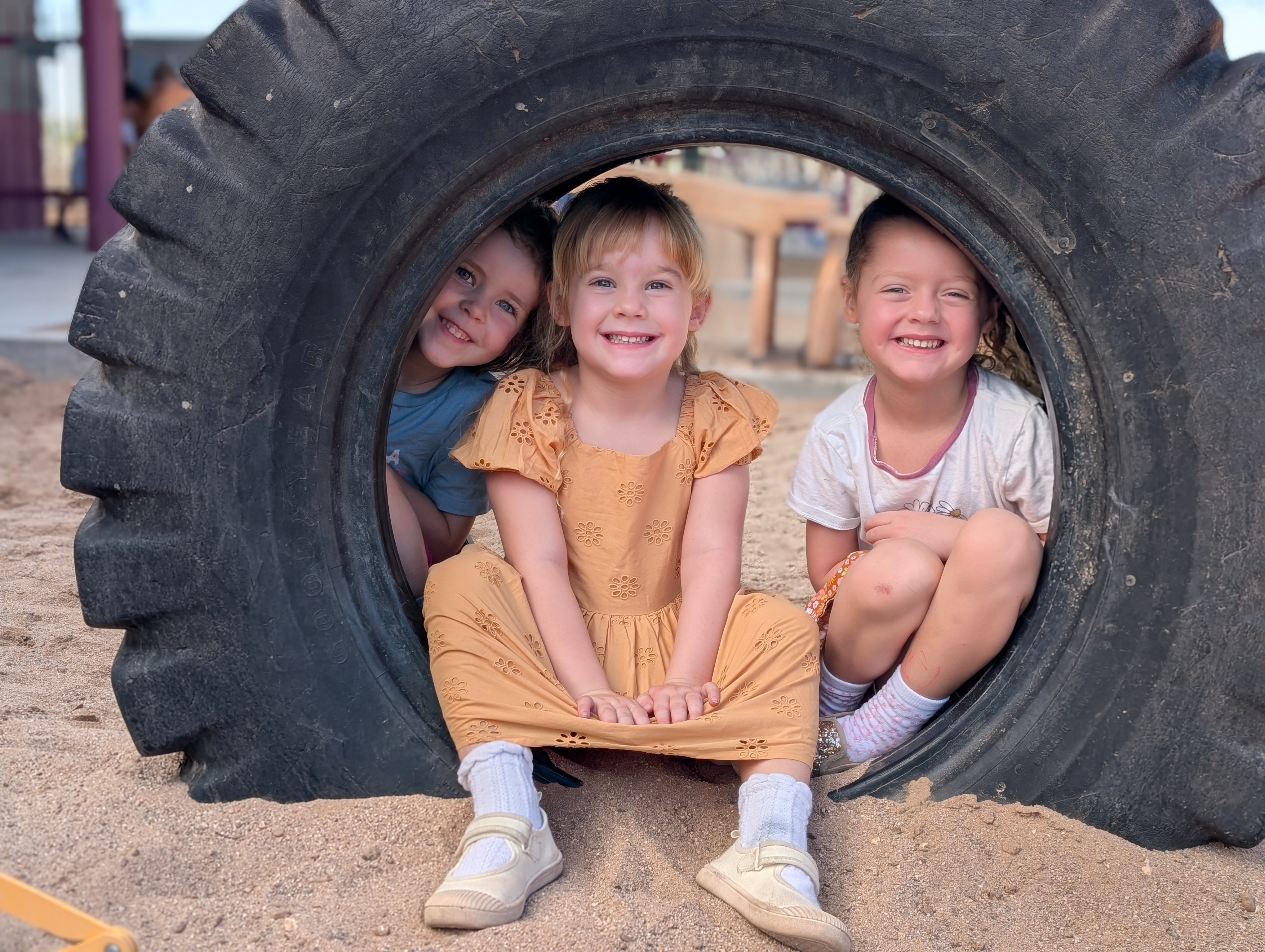 Three young girls are sitting inside a large tire, smiling at the camera. Two are wearing dresses, and one is wearing a blue shirt.