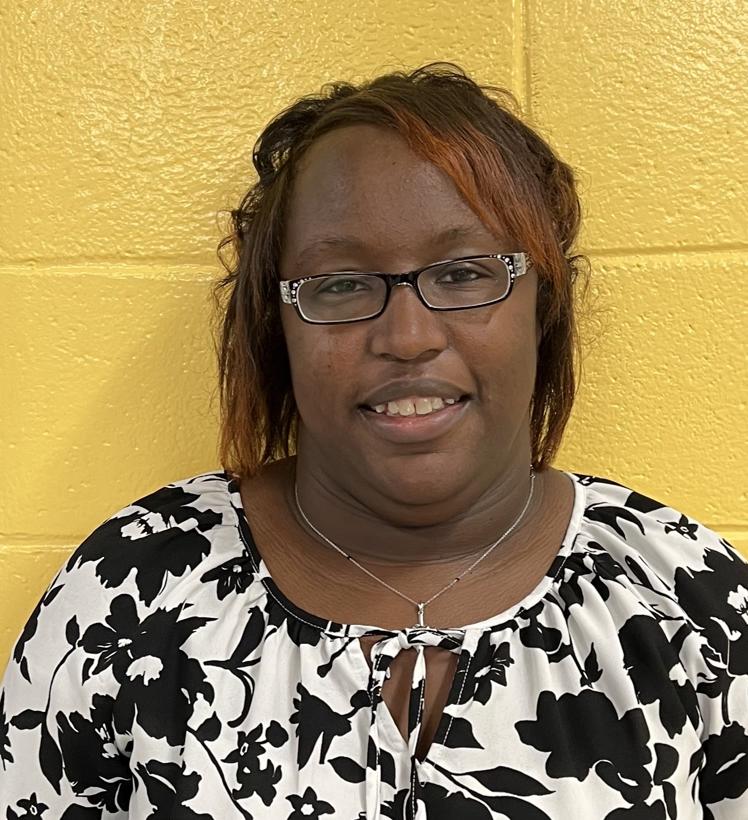 1. A woman in glasses and a floral shirt poses in front of a vibrant yellow wall, showcasing a cheerful and stylish appearance.  2. A woman wearing glasses and a floral shirt stands against a bright yellow wall, exuding a lively and fashionable vibe.  3. A woman with glasses and a floral shirt stands before a sunny yellow wall, radiating a sense of joy and style.