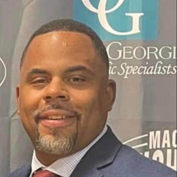 A man in glasses wearing a blue polo shirt, smiling and standing against a neutral background.