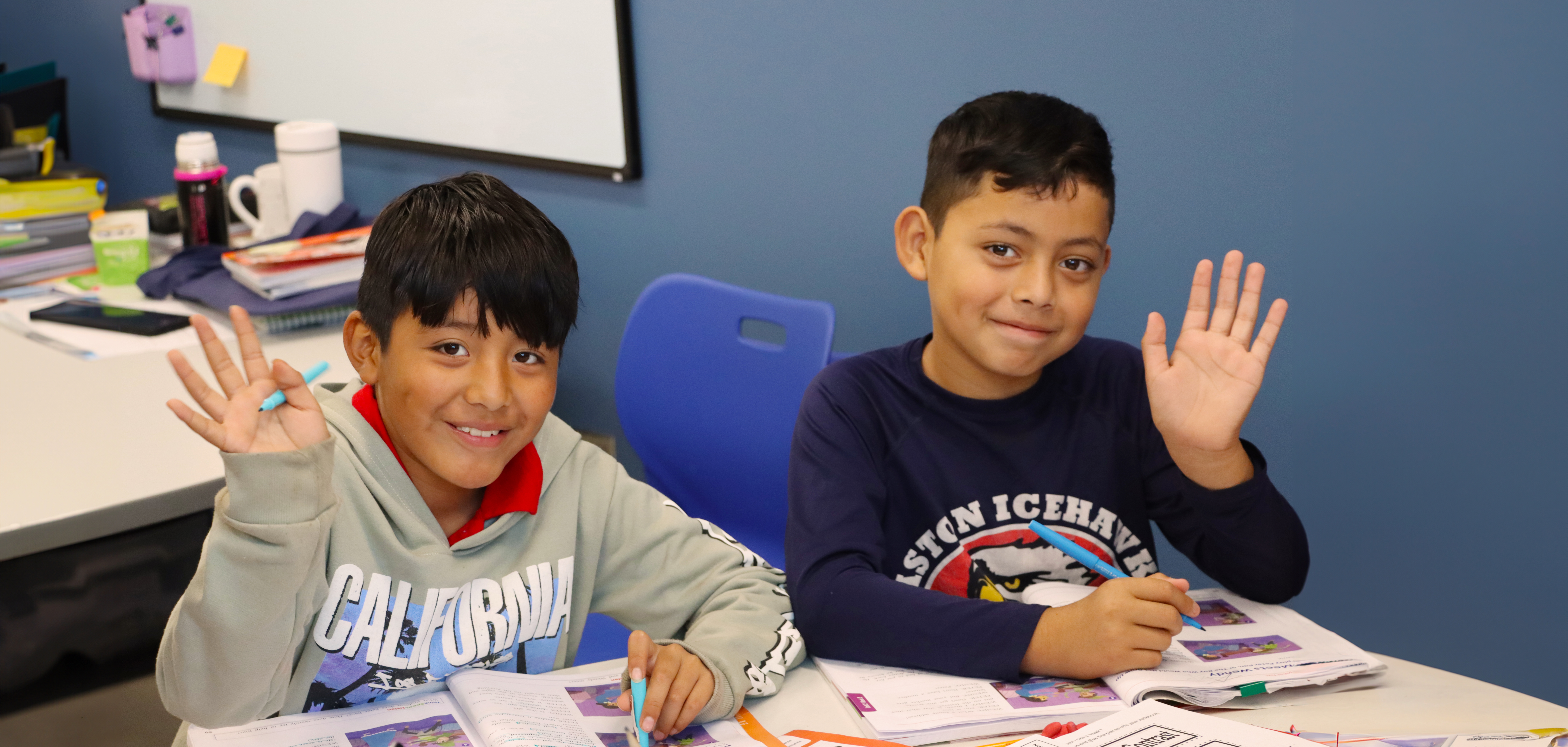 Two boys sitting at a desk waving at the camera
