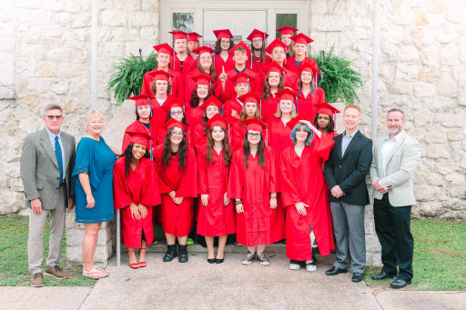 A group of graduates wearing red caps and gowns poses together on steps in front of a stone building, with some adults standing beside them.
