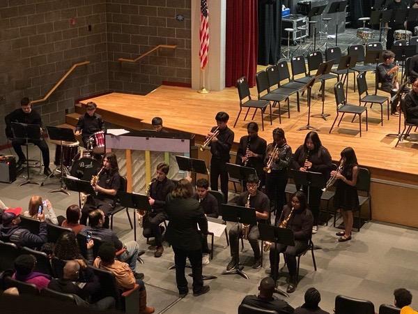 A group of musicians in black attire performs on stage in a school auditorium, with an audience seated in front and empty chairs arranged behind them.