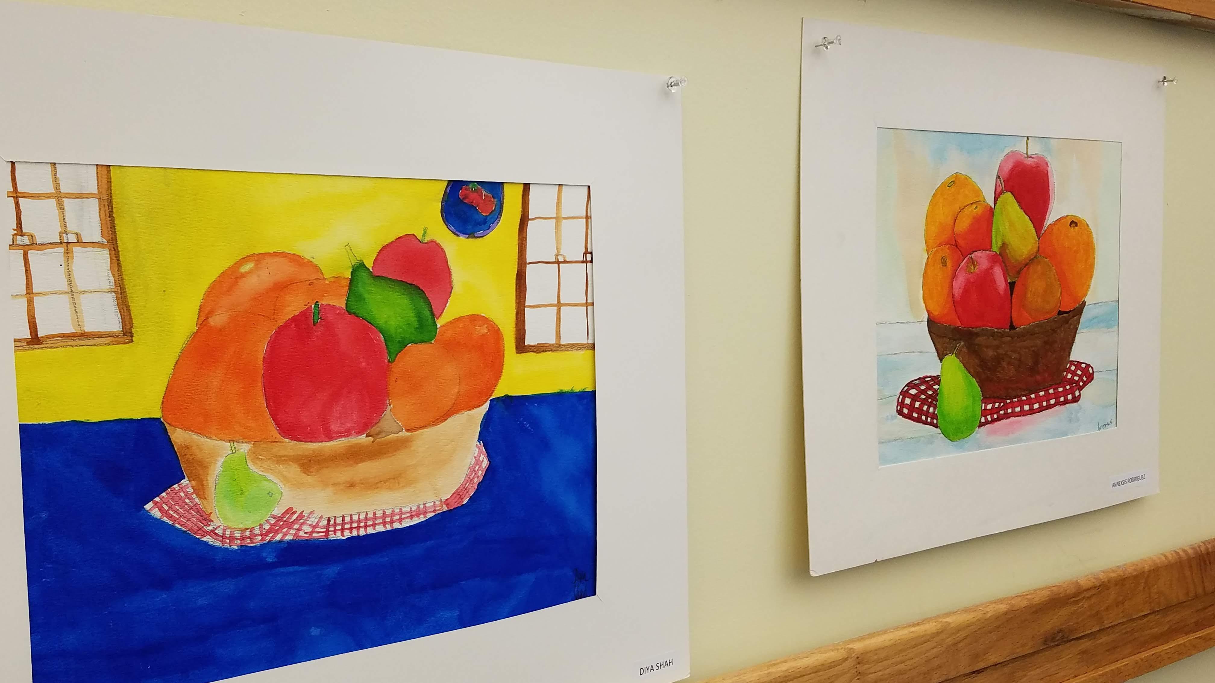 Two colorful paintings of fruit bowls are displayed on a wall, featuring vibrant oranges, apples, and a green pear, with one painting set against a yellow background and the other against a blue background.
