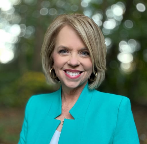 A smiling woman with short blonde hair, wearing a turquoise blazer, stands outdoors with a blurred natural background.