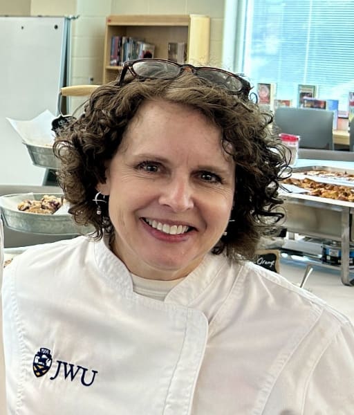 A smiling woman with curly hair wearing a white chef's coat with the initials 'JWU' on it, standing in a kitchen setting with food in the background.