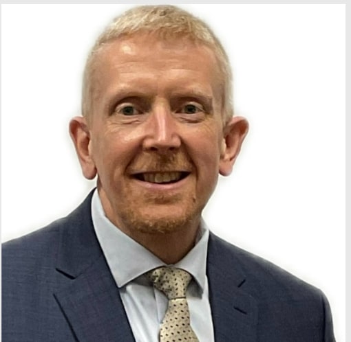 A smiling man with short blonde hair, wearing a dark suit and a light-colored tie, poses against a plain white background.