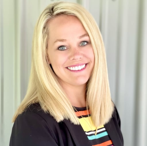 A smiling woman with long blonde hair, wearing a black blazer over a colorful striped shirt, stands in front of a light-colored background.