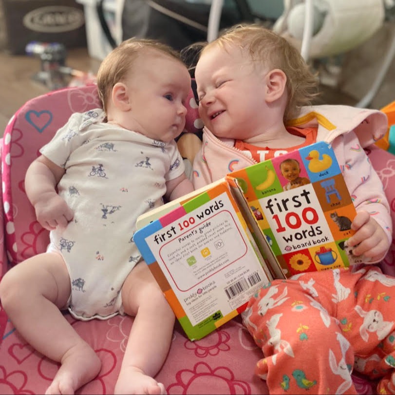A toddler holding a board book "reads" to a baby as the two sit together in a chair