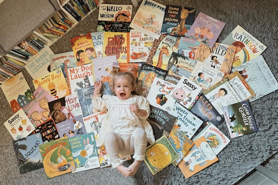 A toddler lying on her back on the floor is surrounded by children's books
