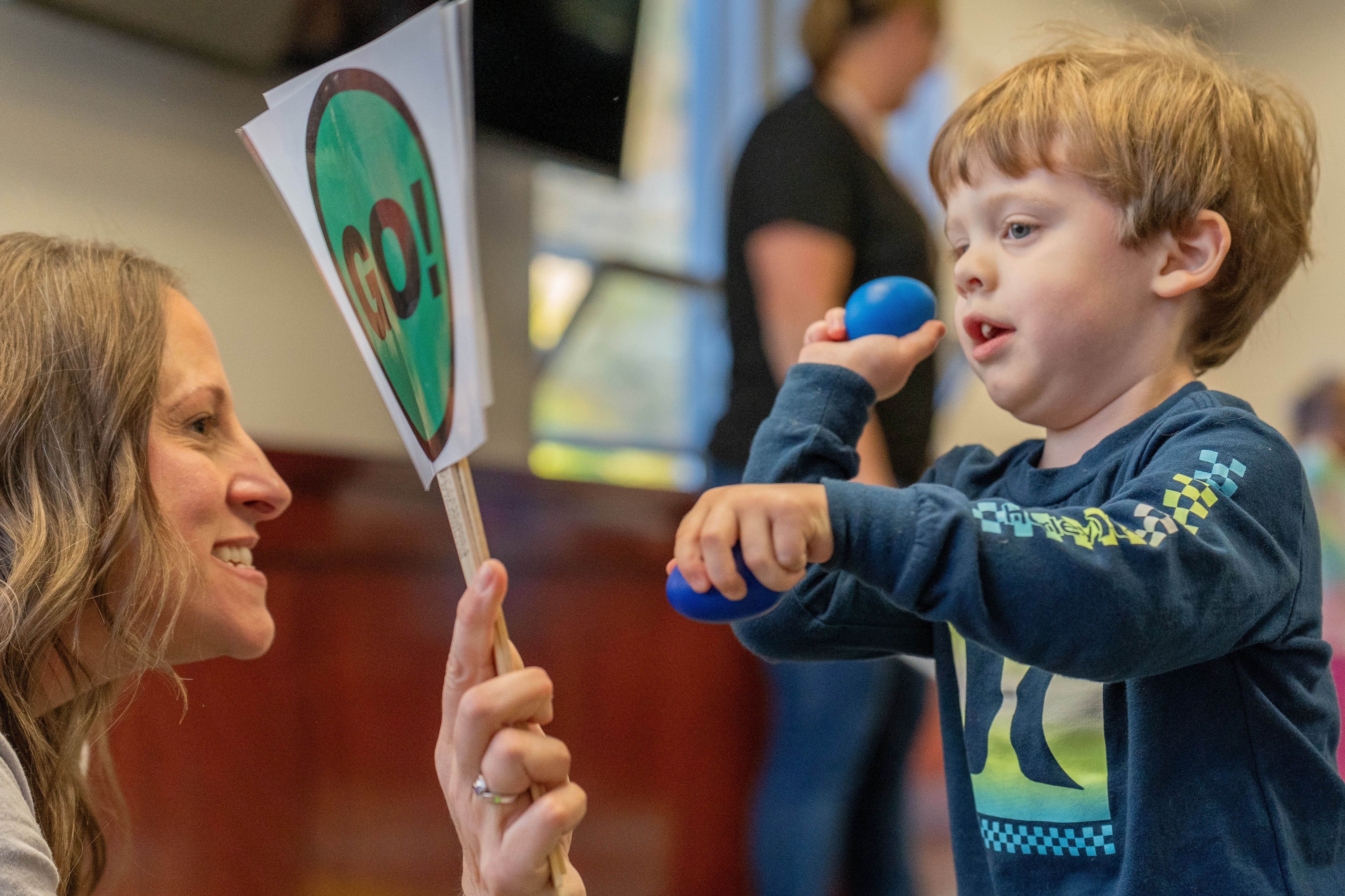 An Early On staff member holds up a paddle reading "Go" while a toddler holding a small blue ball aims for the sign