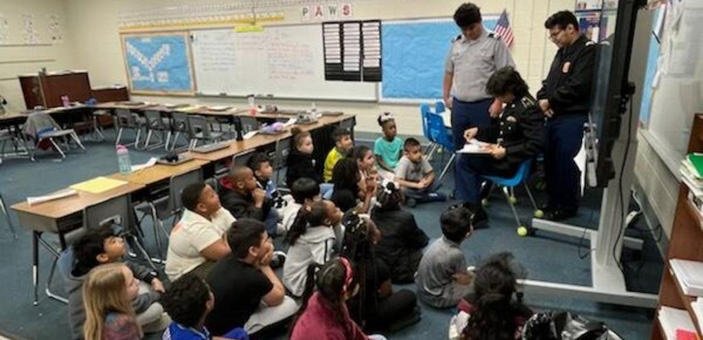 A teacher instructs a group of children seated at desks in a bright classroom, engaged in learning activities.