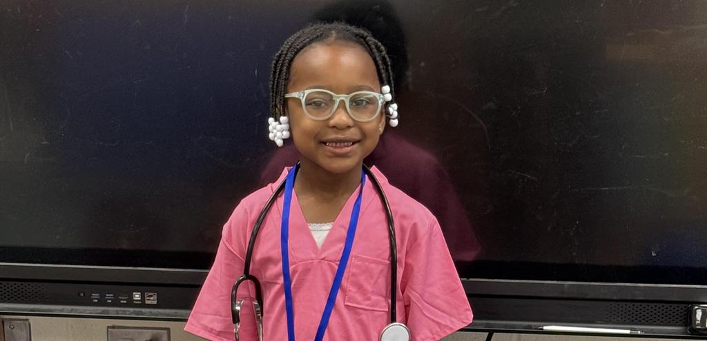 A young girl with glasses wearing a pink shirt, smiling and looking towards the camera in a bright setting.