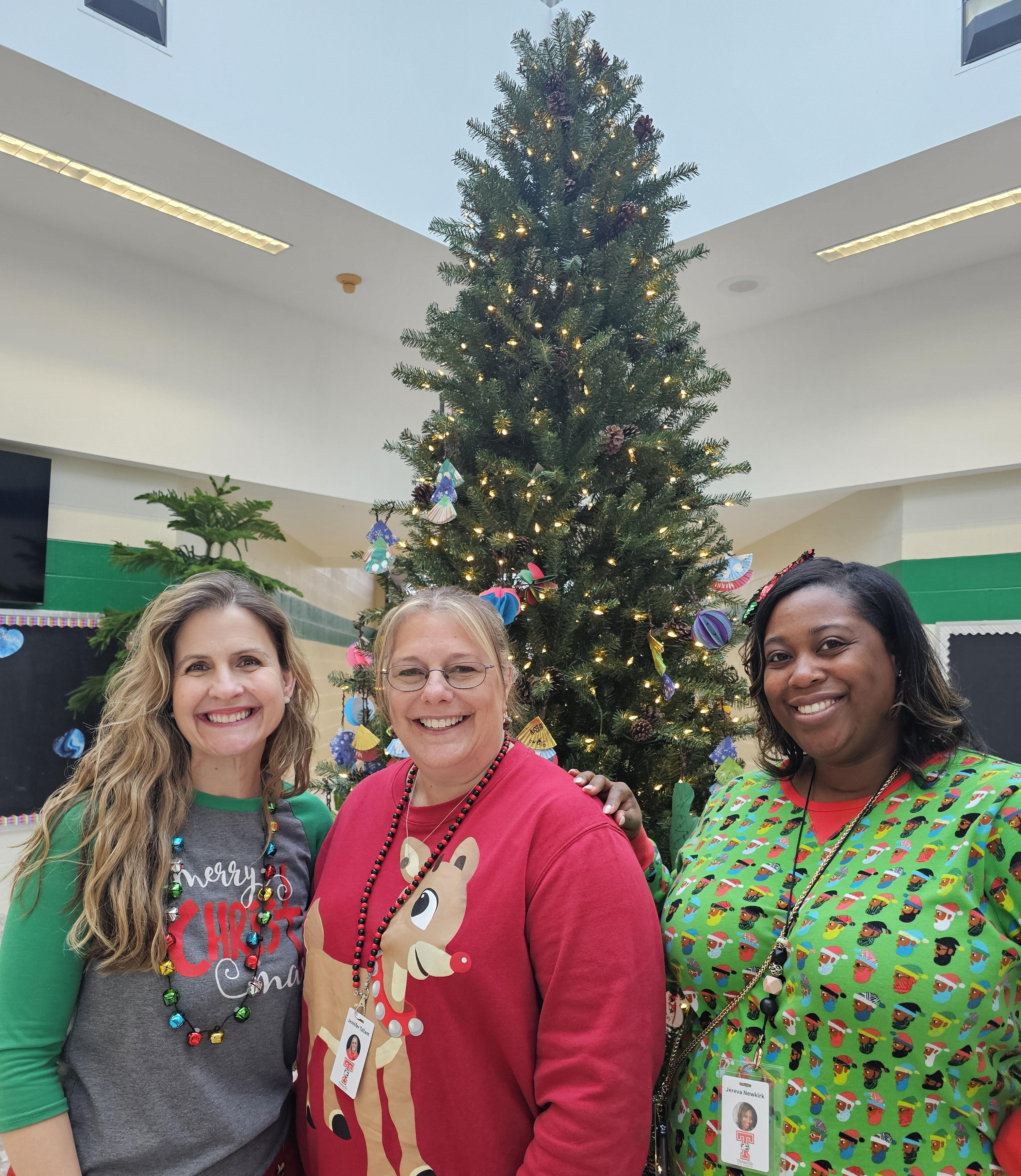 Three women in matching Christmas sweaters pose cheerfully in front of a beautifully decorated Christmas tree.