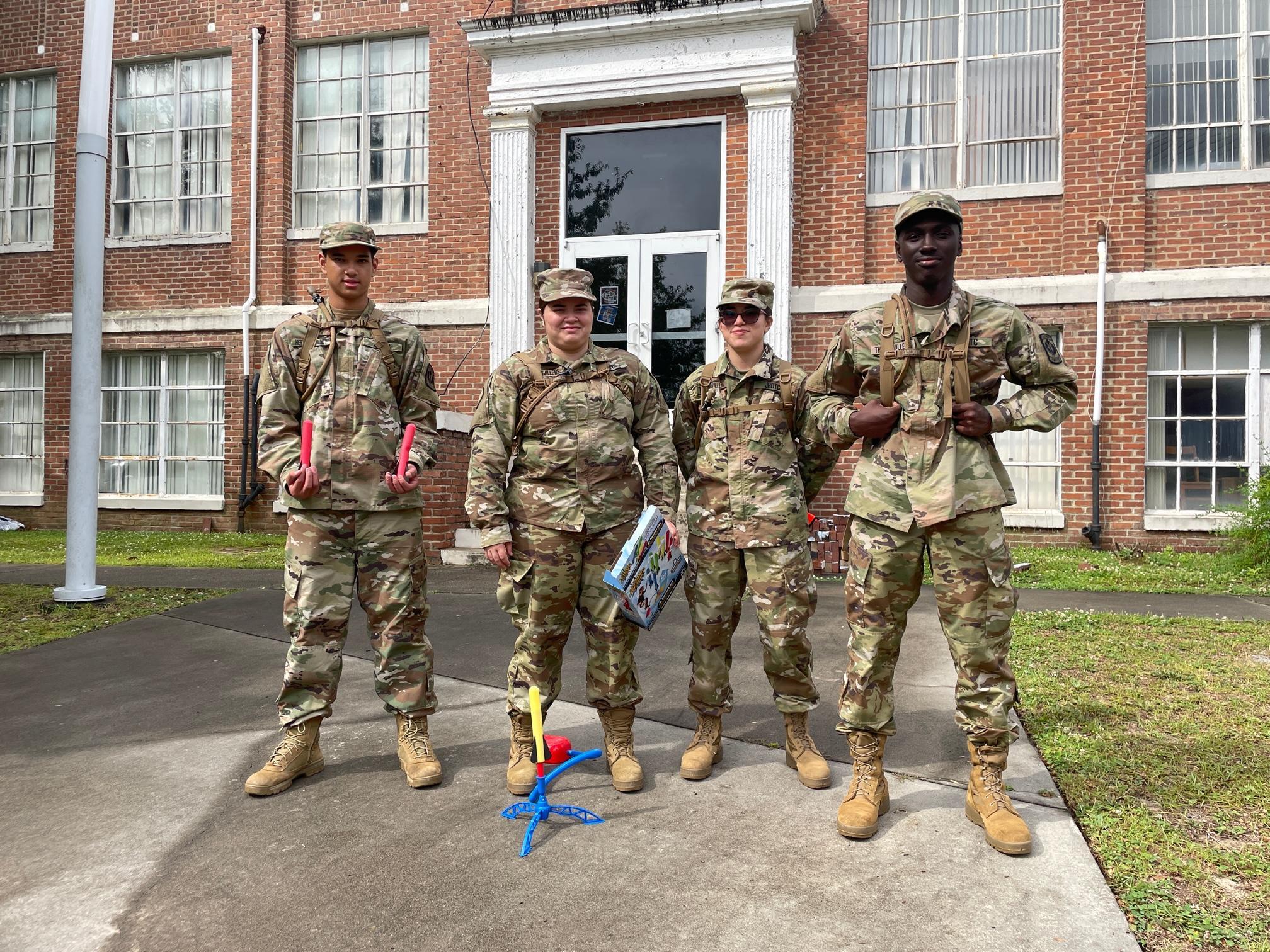 Four soldiers pose confidently in front of a brick building, showcasing camaraderie and strength in their military attire.