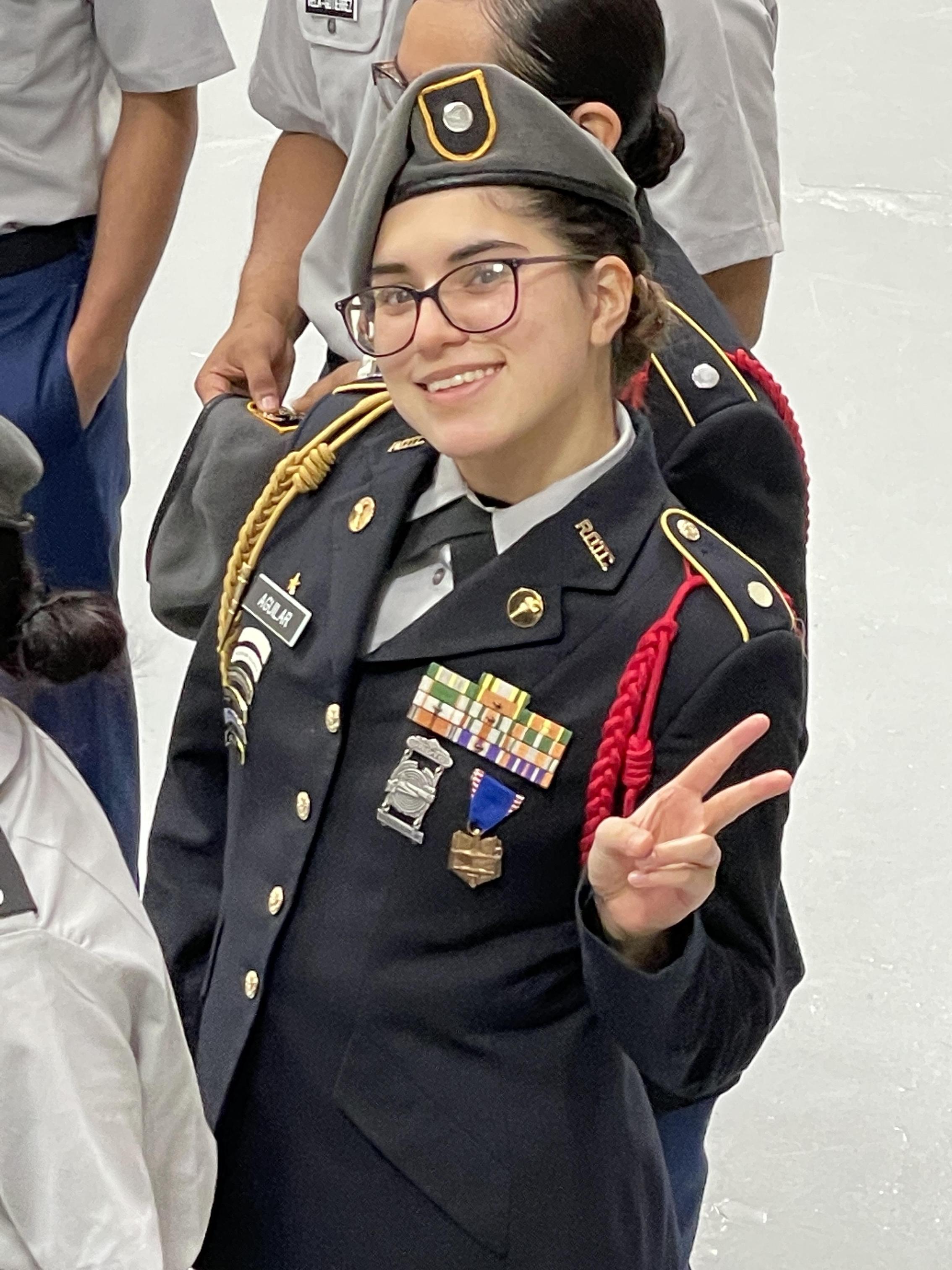 A woman in military uniform stands confidently, smiling and giving the peace sign with her right hand.