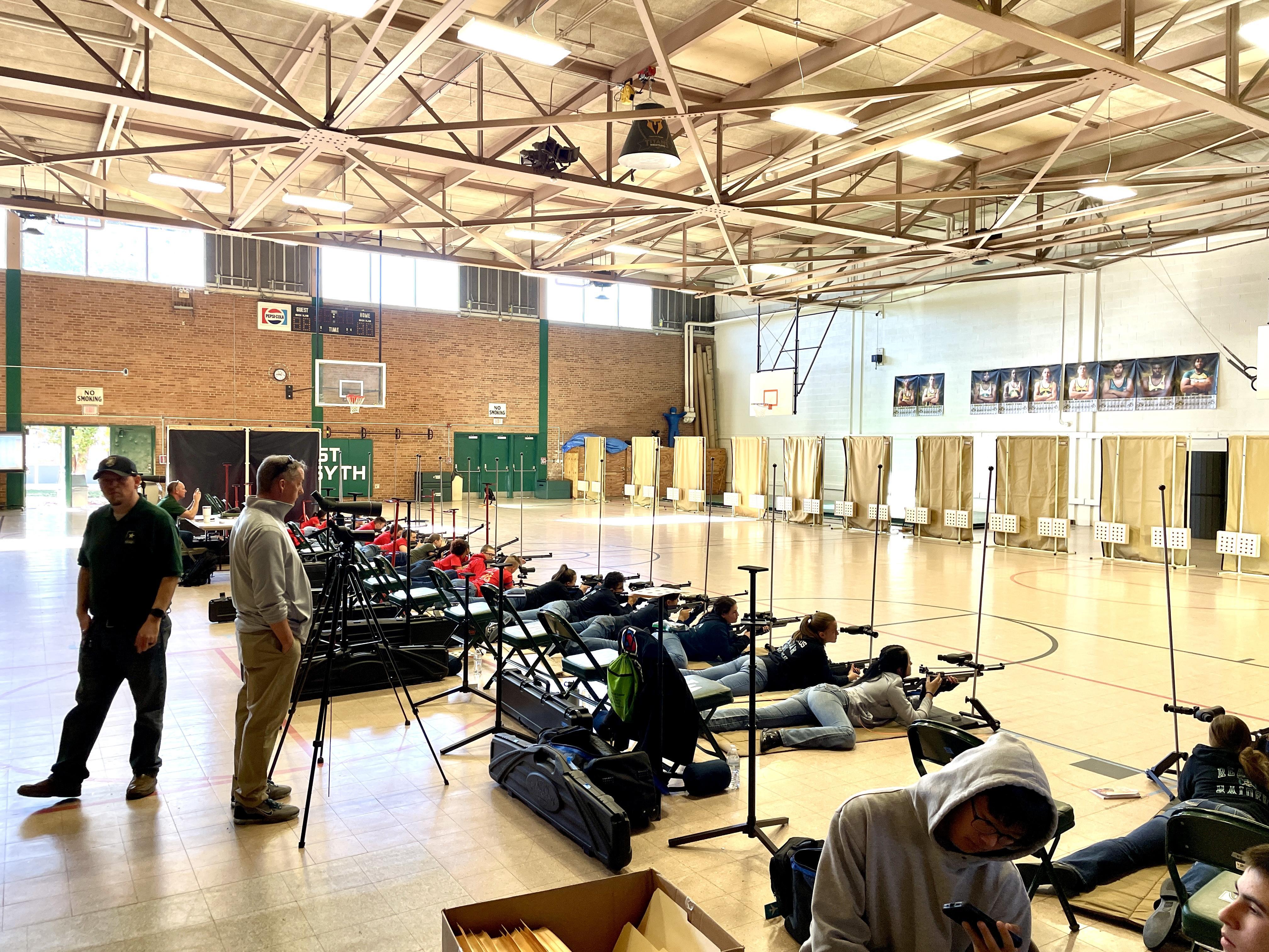 A diverse group of individuals seated in a gym, each holding a firearm, engaged in discussion or training.