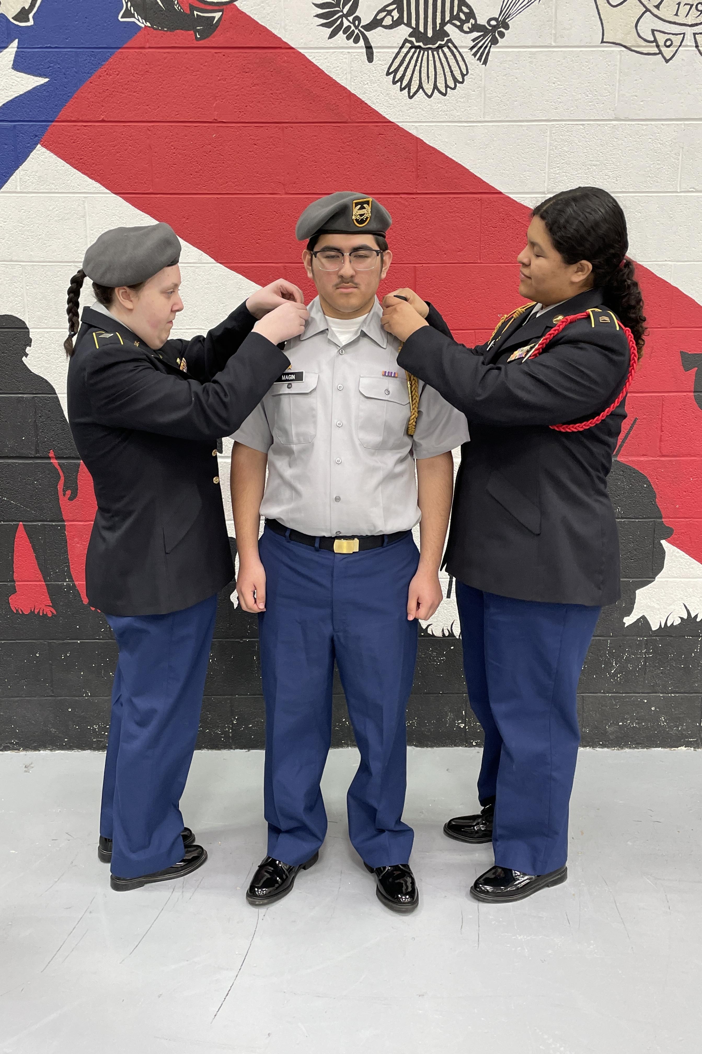 Three women in uniform assist a soldier by adjusting his military attire with precision and care.