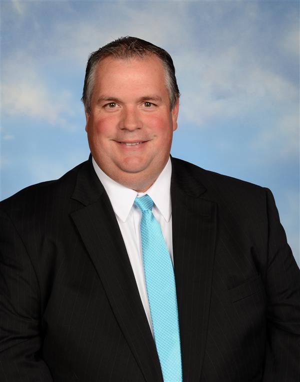 A man in a black suit and light blue tie, seated against a soft blue background, smiling confidently for a portrait.