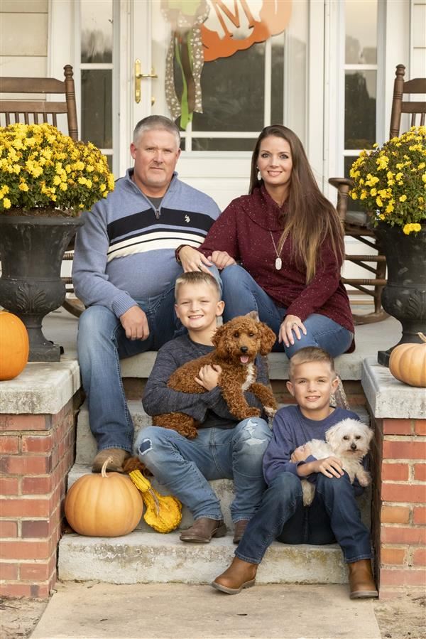 A family of four, including two boys and their parents, sit on the steps of a house decorated with pumpkins and yellow flowers, with the boys holding small dogs.