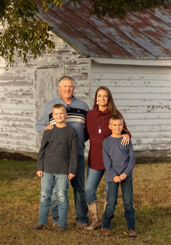 A family of four, consisting of two adults and two children, stands together in front of a weathered white barn with a rusty roof, surrounded by grass.