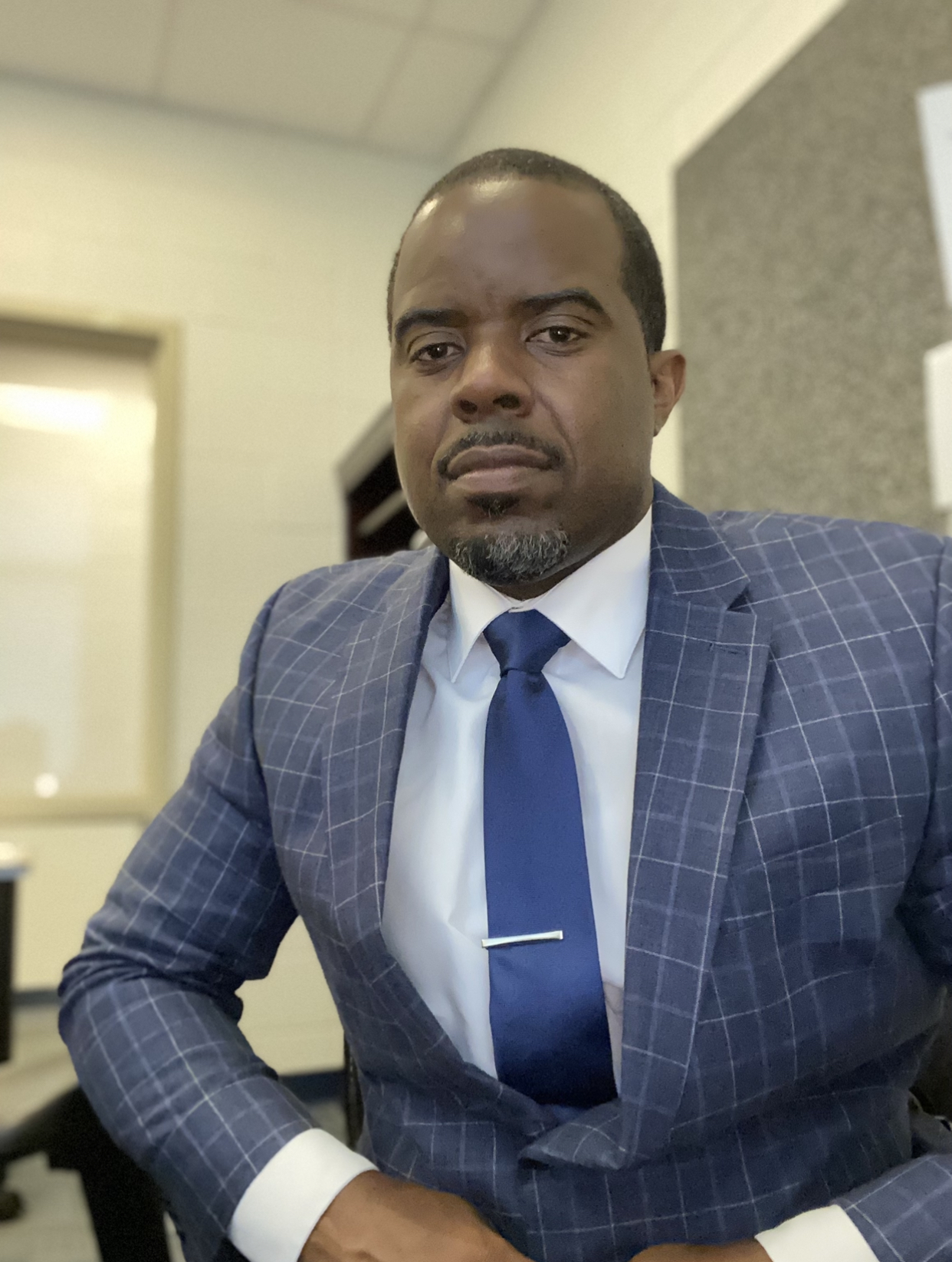A man in a blue checkered suit and tie sits at a desk, looking directly at the camera with a serious expression.