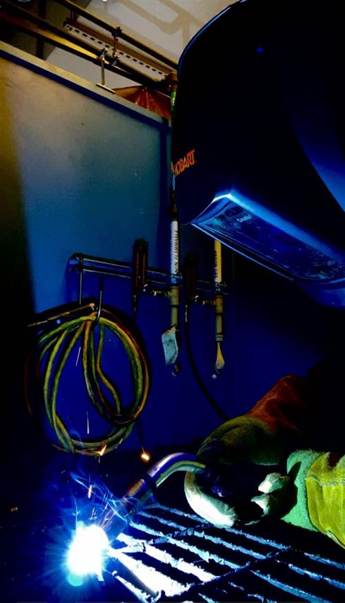 A close-up view of a person welding, with bright sparks and a blue flame illuminating the workspace, surrounded by hoses and equipment.